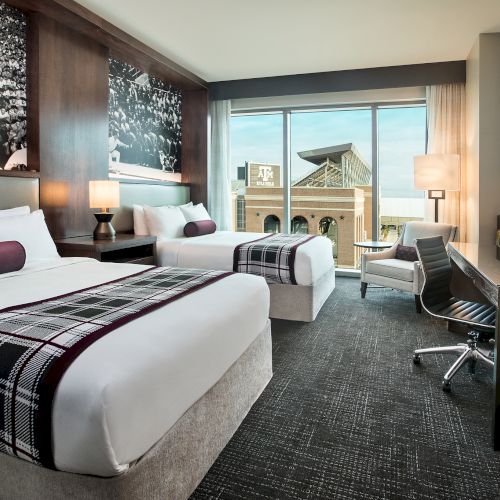 The image shows a modern hotel room with two beds, a large window, a desk, a TV, and stylish decor, featuring a monochrome wall art.