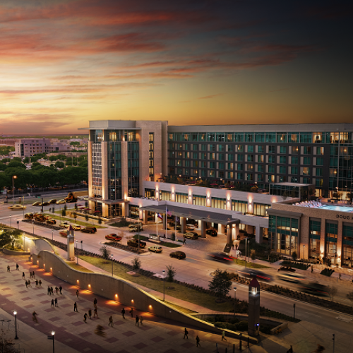 A large hotel complex at sunset with surrounding roads, trees, and a plaza featuring walking paths and sculptures.