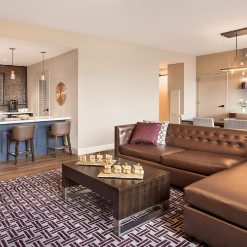 A modern living room with a brown leather sofa, patterned rug, coffee table, open kitchen, bar stools, and decorative lighting.