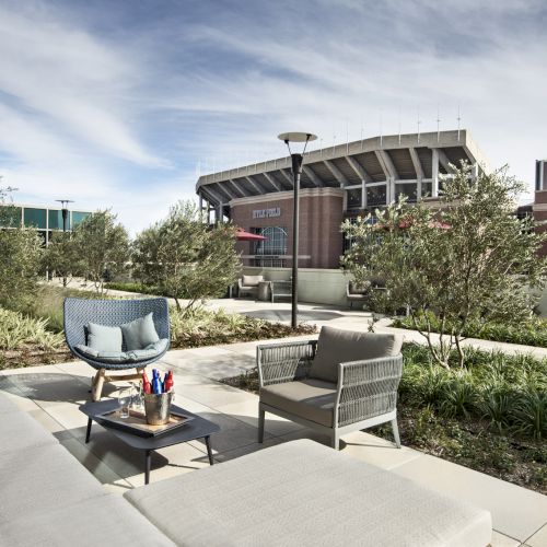 The image shows outdoor seating with modern furniture on a patio, overlooking a stadium surrounded by greenery and trees under a blue sky.