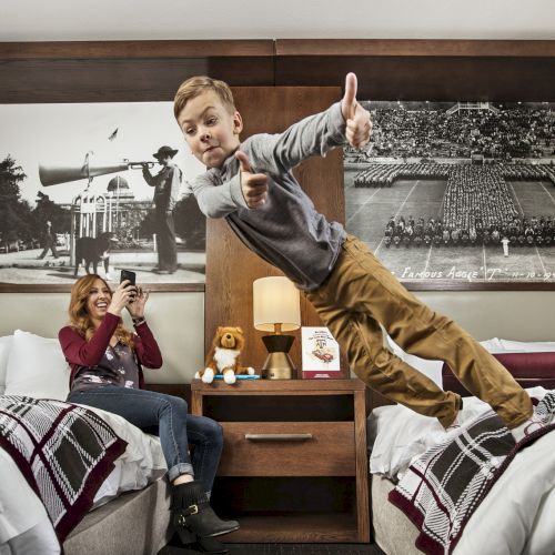A child playfully jumps between beds in a hotel room as a woman sits nearby, engrossed in her phone, with vintage photos decorating the wall.