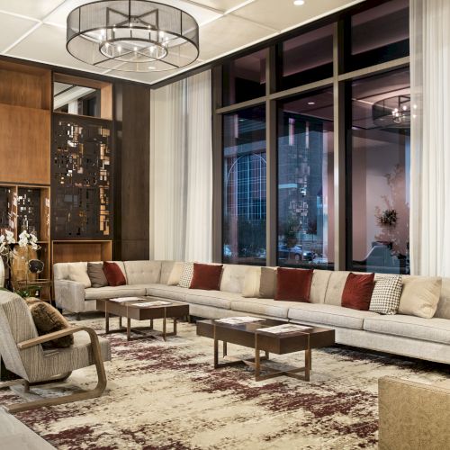 The image shows a modern lounge with a large sectional sofa, armchairs, coffee tables, and decorative shelves, set on a patterned rug.