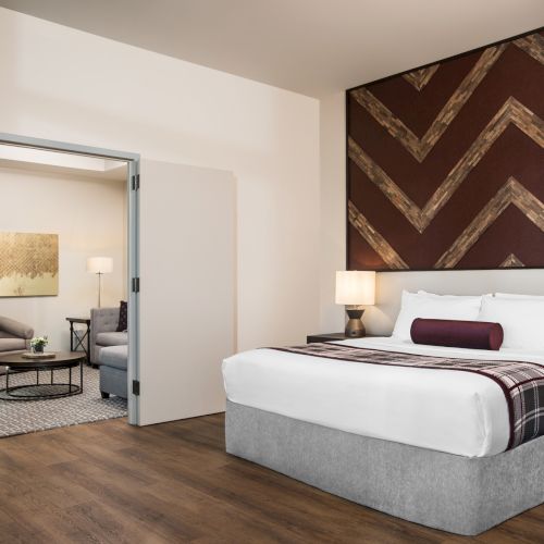 A hotel room with a neatly made bed, decorative headboard, seating area with sofa and chairs, and wooden flooring.