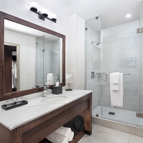 The image shows a modern bathroom with a large mirror, a wooden vanity, a sink, and a glass-enclosed shower with a towel hanging inside.