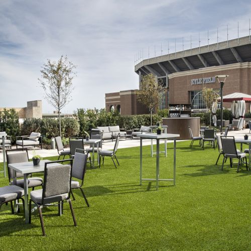Outdoor seating area on a grass lawn with tables and chairs in front of a building that resembles a stadium.