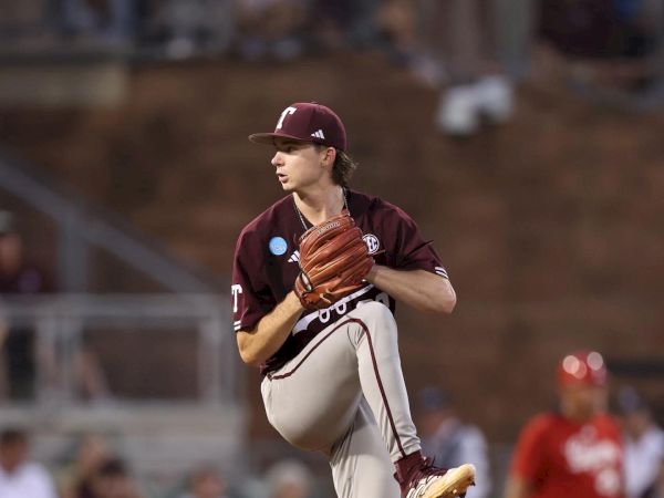 An Aggie baseball player in a maroon uniform is pitching on the mound, with a runner on base and spectators in the background.