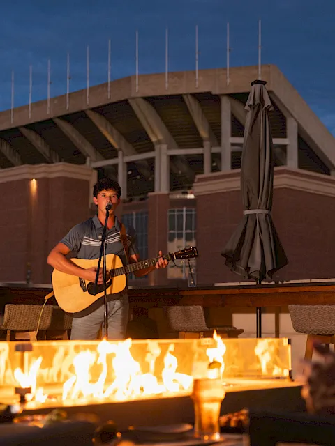 A guitarist sings by a crackling bonfire at an outdoor event under a stadium-style roof.