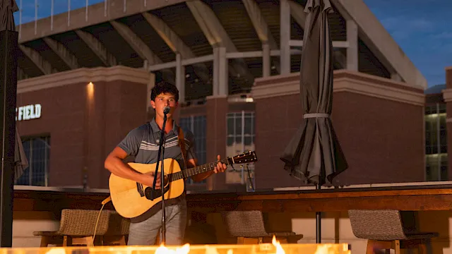 A guitarist sings by a crackling bonfire at an outdoor event under a stadium-style roof.