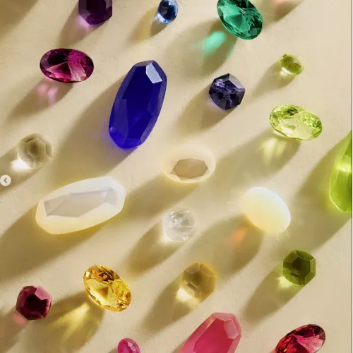 A spread of colorful faceted gemstones&mdash;purple, blue, green, yellow, and red&mdash;scattered on a light background.
