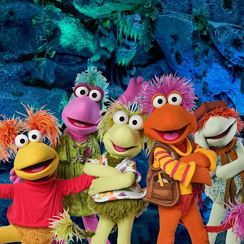 Five colorful Sesame Street-style puppet characters sing together in an underwater blue cave, wearing bright wigs and costumes.