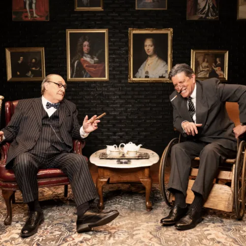 Two men in a cozy room play-act tea-time: one in a wheelchair pours, the other sits chatting beside a teapot and cups.