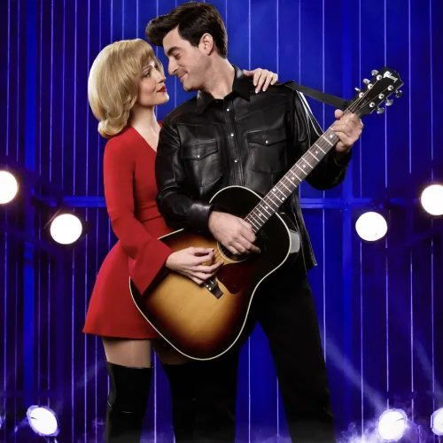 A guitarist in a black jacket serenades a woman in a red dress on a blue stage with spotlights and fog.