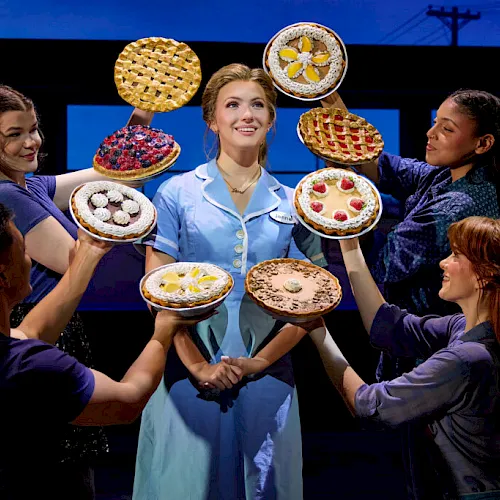 A contestant smiles center stage as helpers surround her with colorful pies and waffles on trays in a blue studio, everyone grins.