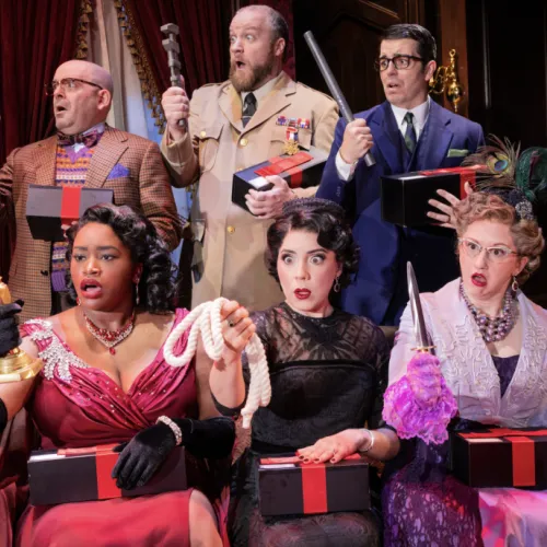 A comedic troupe in vintage costumes on a theater stage holds prop guns and gifts in red-and-gold boxes.