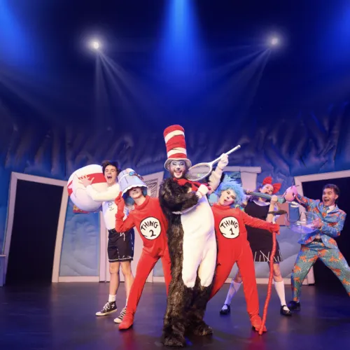 A circus troupe in red-and-white outfits dances under blue spotlights, with a clown holding a tennis racket and a bear-costumed performer.