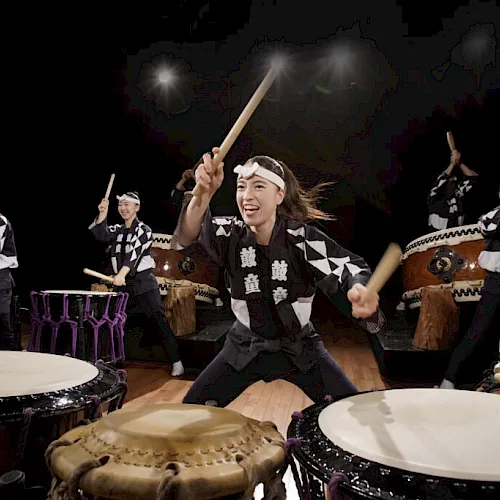 A group of drummers in matching black-and-white outfits performs with sticks and large taiko drums under bright stage lights.