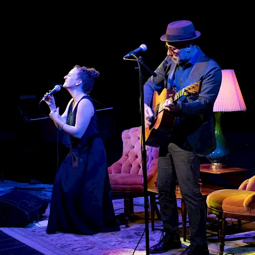 A live music performance on stage: a singer at a microphone and a guitarist in a hat playing beside him, under purple lights.