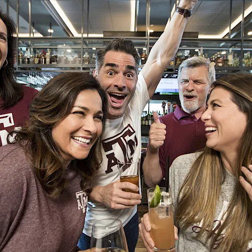 Friends in matching maroon cheer in a bar, holding drinks and laughing as one person raises an arm&mdash;game night energy!