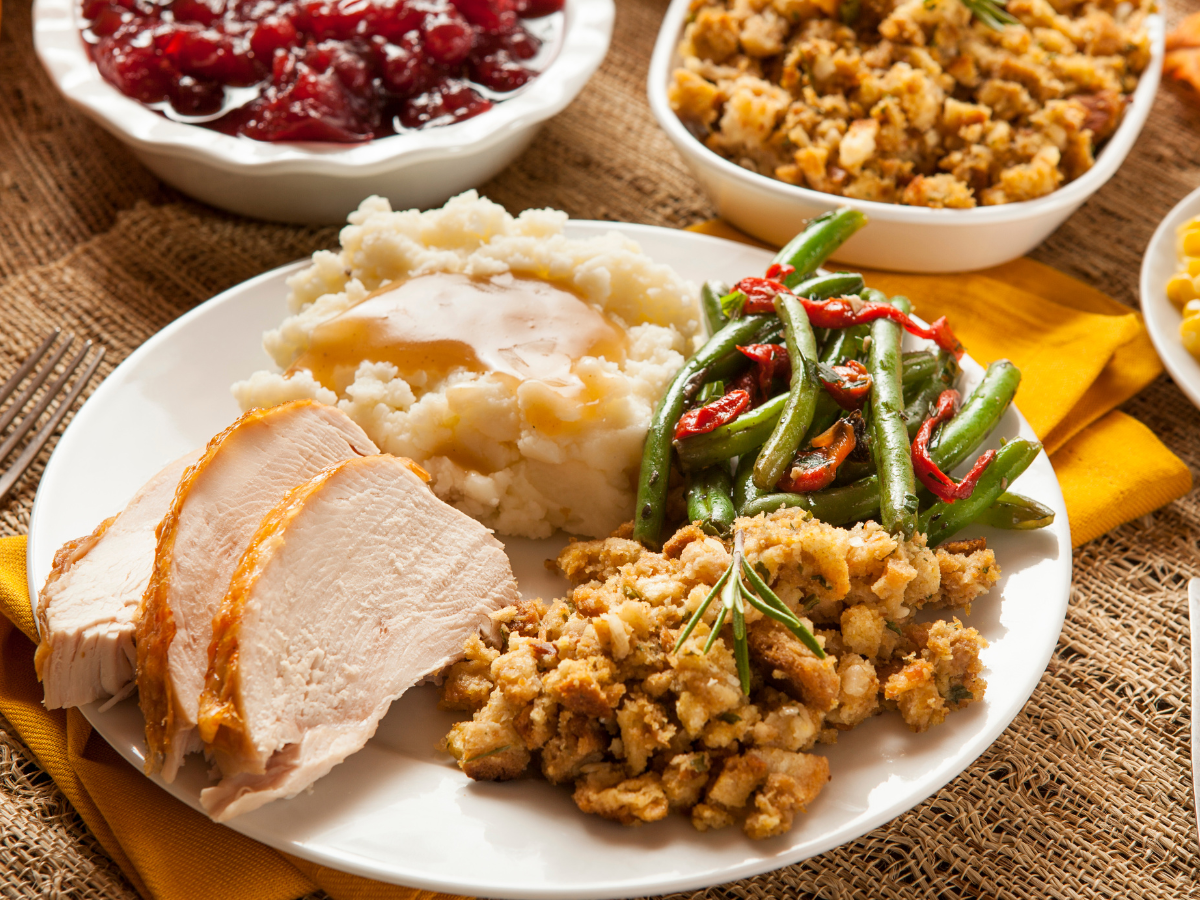 A plate of Thanksgiving food featuring slices of turkey, mashed potatoes with gravy, green beans, and stuffing, surrounded by bowls of cranberry sauce and more stuffing.
