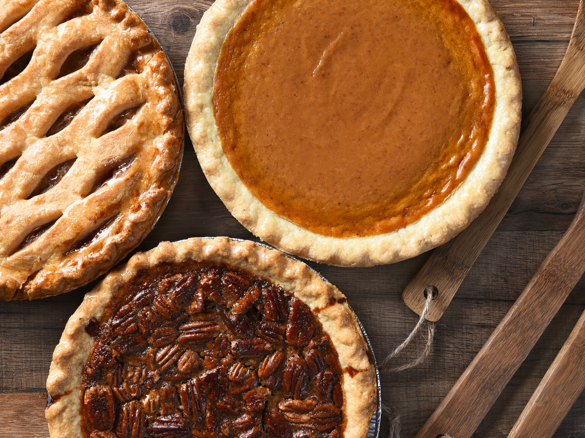 The image shows three pies: an apple pie with a lattice crust, a pumpkin pie, and a pecan pie, placed on a wooden table with utensils nearby.