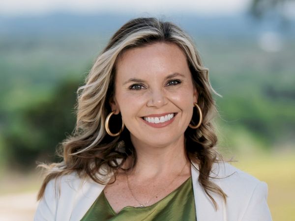 A smiling woman wearing a green top and white jacket poses outdoors with a blurred natural background.