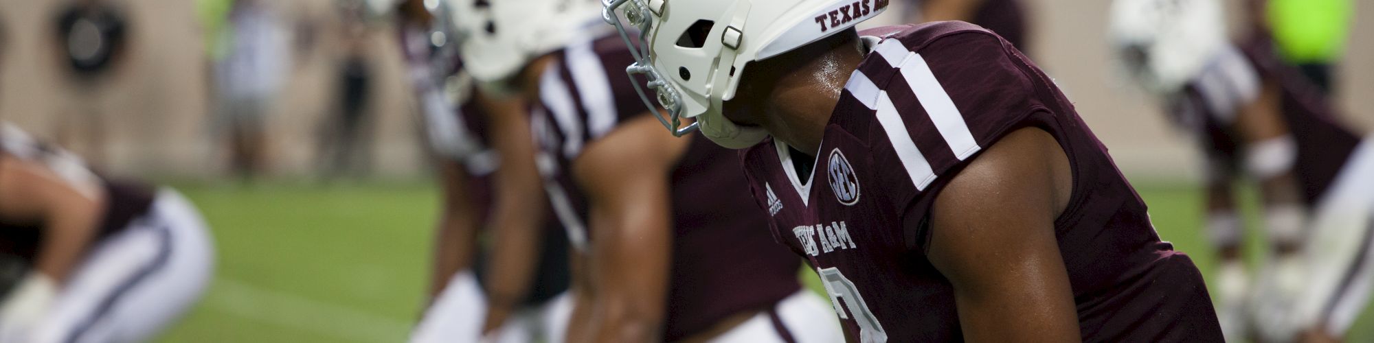 This image depicts a football team in maroon and white uniforms lined up on the field, focused and ready for a play, during a game.