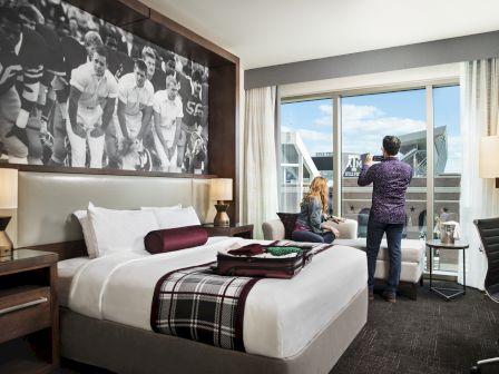 A hotel room with a large bed, a black-and-white football photo on the wall, and two people looking out the window towards a stadium.