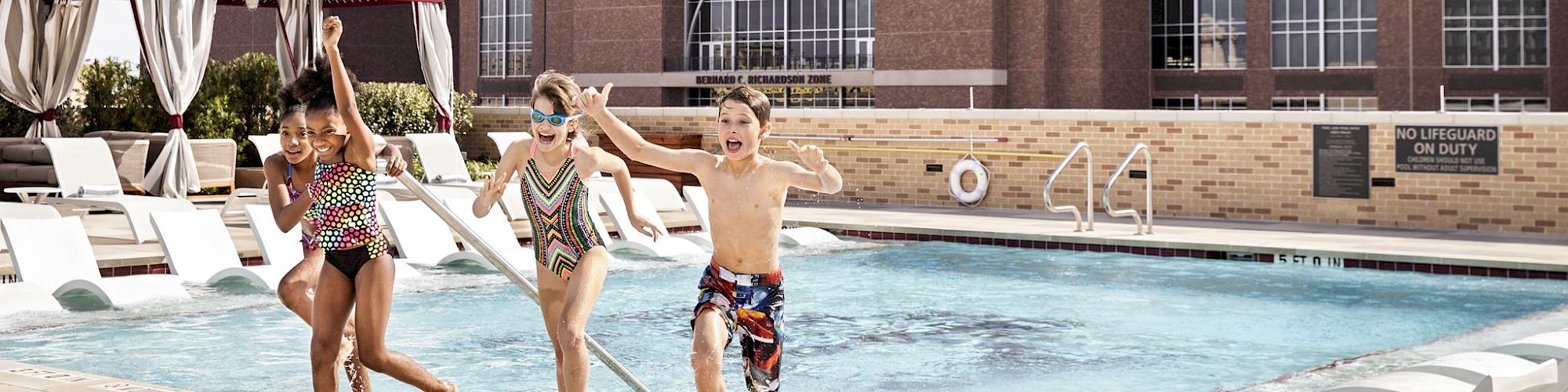 Four children in swimwear are jumping into a pool in front of a building with a sign that reads "KYLE FIELD". The pool area has loungers and cabanas.