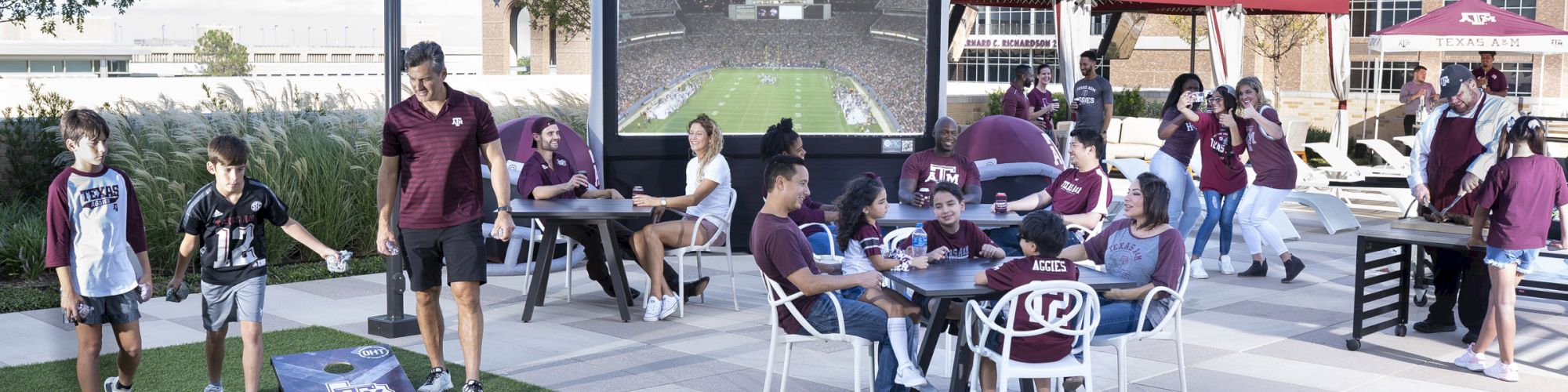 Hullaballoo Gameday Event on the pool deck with a screen showing the Aggie game on a projector, kids playing cornhole, and people enjoying conversation at tables