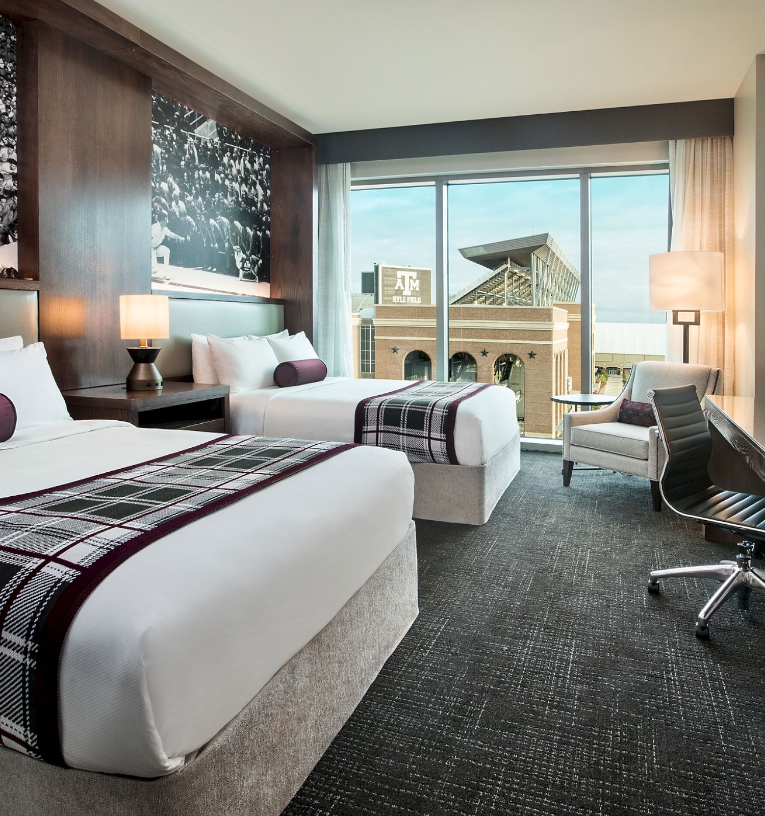 A hotel room with two double beds, a desk with a chair, a flatscreen TV, and large window showing an outdoor view; modern and cozy decor.