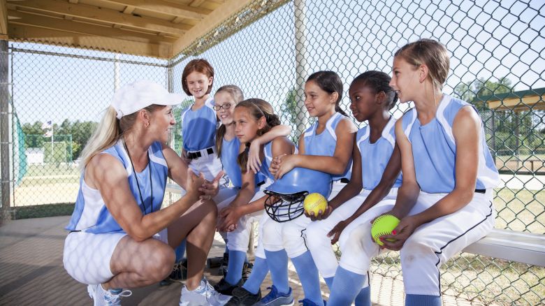 A coach is talking to a group of young softball players in a dugout, all in blue uniforms, holding softball equipment and looking attentive.