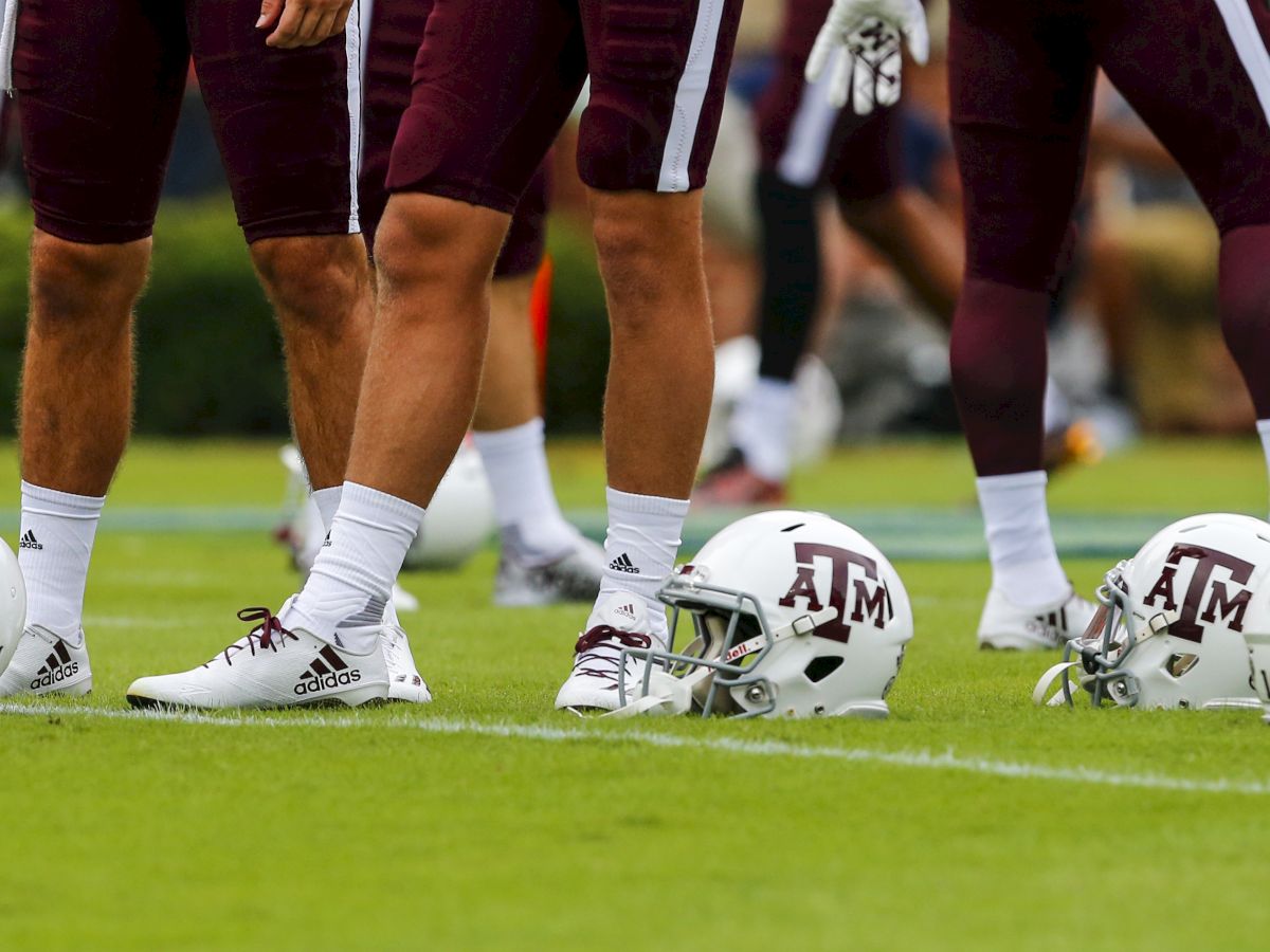 Football players in maroon uniforms stand on a field; white helmets with "A&M" logos are placed near their feet.