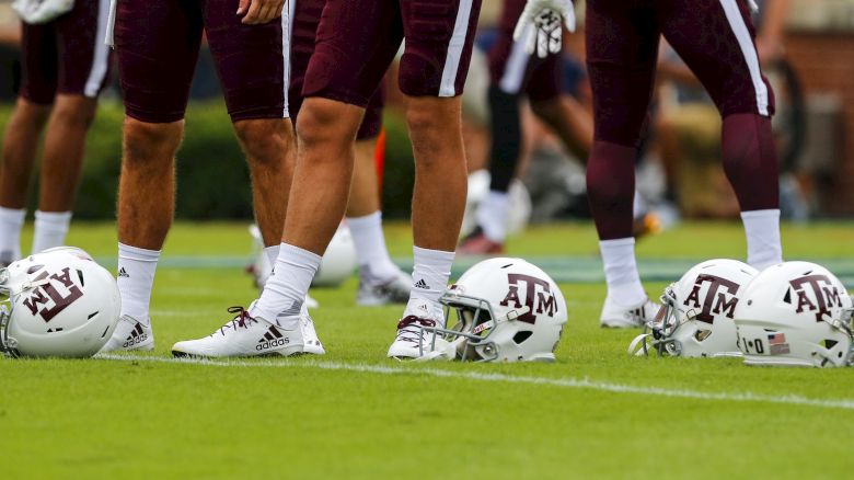 Football players in maroon uniforms stand on a field; white helmets with "A&M" logos are placed near their feet.