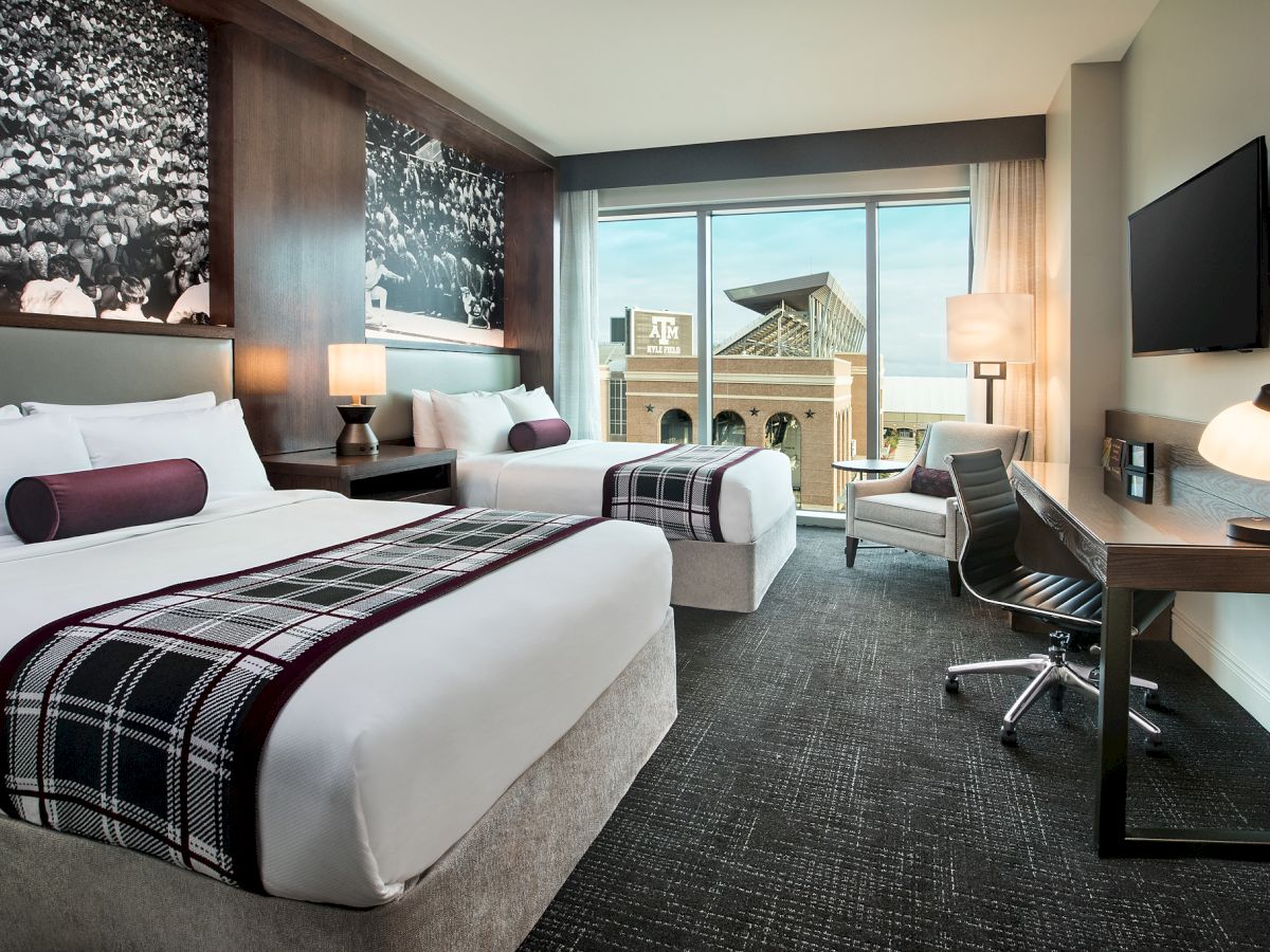 A modern hotel room with two beds, a desk, a chair, a wall-mounted TV, lamps, and large window offering a scenic view.