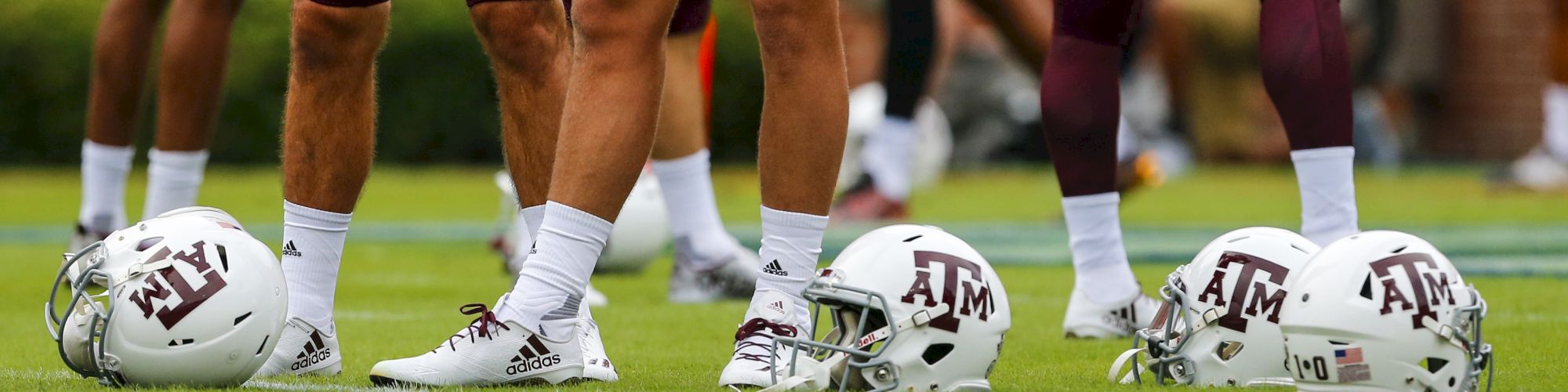 Football players standing on a field with their white helmets on the ground, marked with "A&M" logos on them.