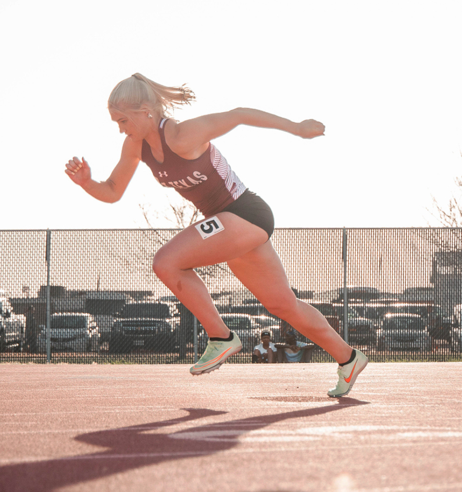 A sprinter wearing a maroon and white uniform launches off the starting block on an outdoor track, with cars and trees in the background.