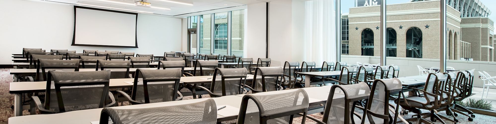 An empty conference room with rows of chairs and desks, a large screen at the front, and large windows offering a city view.