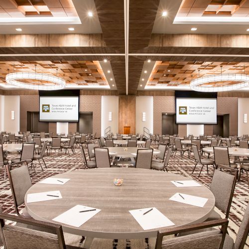 The image shows a spacious conference room with round tables, chairs, screens, and chandeliers, ready for an event or meeting.