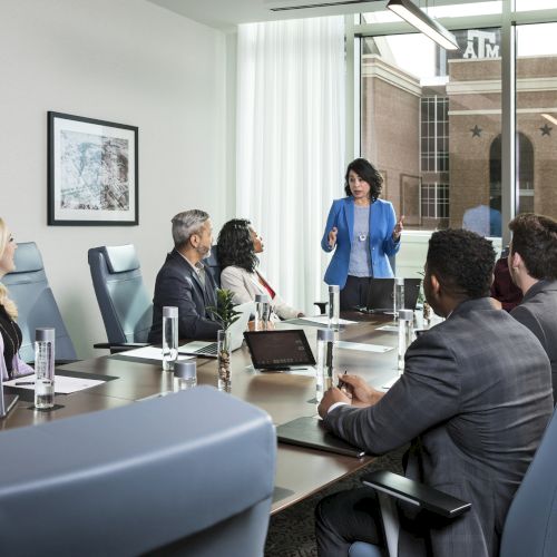 A business meeting with eight people around a conference table, one standing and speaking, with laptops and water glasses present.