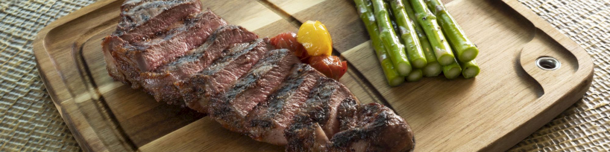 A wooden plate with a grilled steak, asparagus, and a small red pepper. The plate is placed on a textured woven surface.