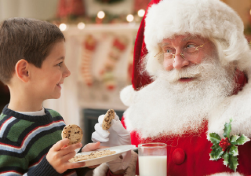 A child offers cookies to Santa Claus, who holds a glass of milk, in a festive setting with Christmas decorations in the background.