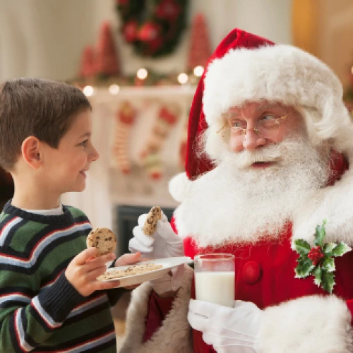 A child offers cookies to Santa Claus, who holds a glass of milk, in a festive setting with Christmas decorations in the background.