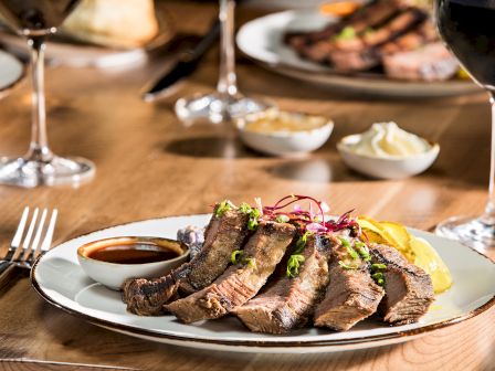 Slices of grilled steak garnished with herbs, lemon, and sauce, served on a plate with wine glasses in the background, garnishing on top.