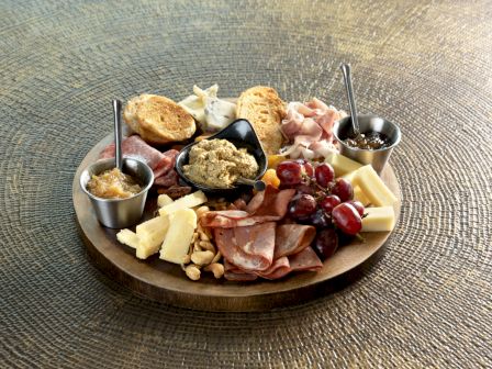 A charcuterie board with meats, cheeses, grapes, nuts, spreads, and bread slices on a textured surface.