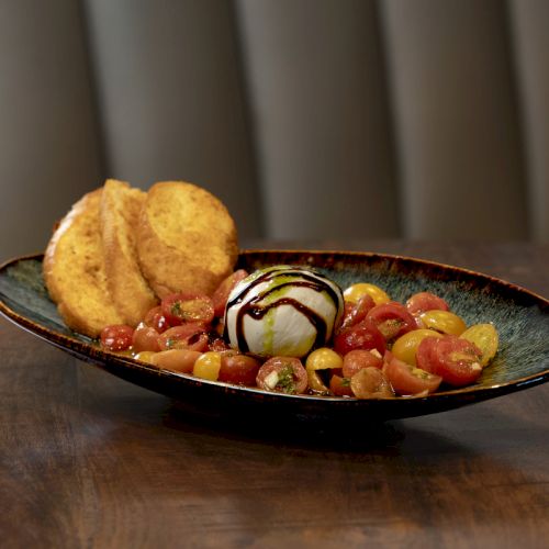 A dish with sliced tomatoes, burrata cheese drizzled with balsamic glaze, and toasted bread slices on the side, served on a dark plate.