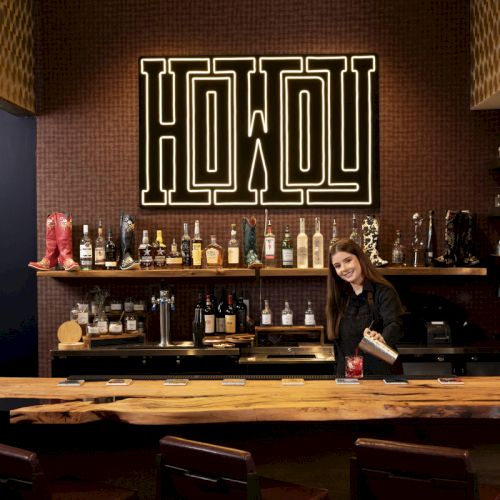 A bartender stands behind a wooden bar counter in a stylish setting with a "HOWDY" sign and shelves stocked with various bottles and glasses.