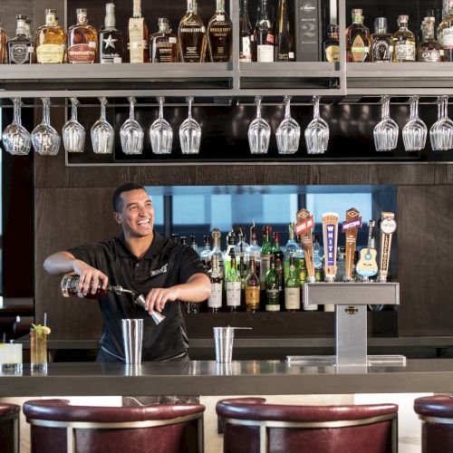 A bartender is preparing drinks behind a bar with a variety of bottles and beer taps, under hanging glasses, in a well-lit lounge.