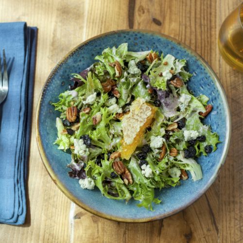 A salad with mixed greens, blue cheese, pecans, and a citrus slice, alongside a drink, knife, and fork set on a blue napkin.