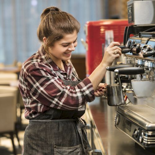 A barista in a plaid shirt is using an espresso machine in a cafe, smiling while steaming milk.