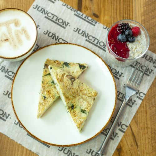 The image shows a cappuccino, two slices of frittata on a plate, and a chia pudding with berries, on a wooden table with branded paper.